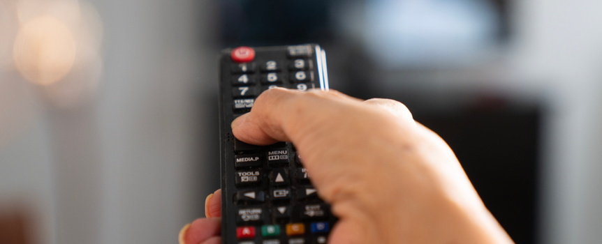 Na zdjęciu widać rękę trzymającą pilot telewizyjny, wskazującą na ekran telewizora.