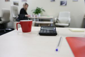 Na biurku kawa w czerwonym kubku, kalkulator, długopis i notatnik. W tle osoba przy stoliku.
