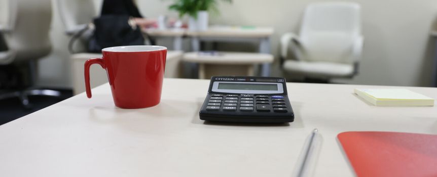 Na biurku kawa w czerwonym kubku, kalkulator, długopis i notatnik. W tle osoba przy stoliku.