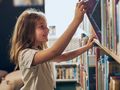 Dziewczynka z długimi włosami uśmiecha się, sięgając po książkę na półce w bibliotece.