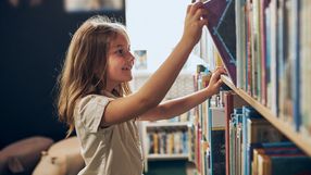 Dziewczynka z długimi włosami uśmiecha się, sięgając po książkę na półce w bibliotece.