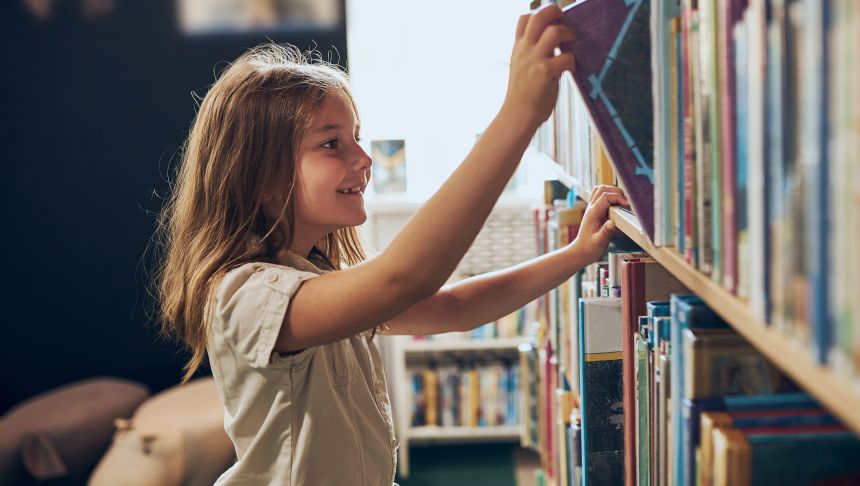 Dziewczynka z długimi włosami uśmiecha się, sięgając po książkę na półce w bibliotece.