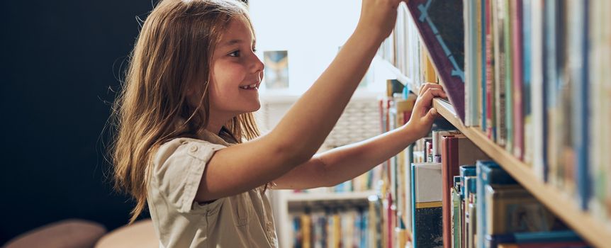 Dziewczynka z długimi włosami uśmiecha się, sięgając po książkę na półce w bibliotece.