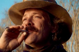 Mężczyzna w kapeluszu pali papierosa, uśmiechając się i spoglądając w stronę nieba.