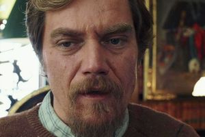 Mężczyzna z brodą, w swetrze, zmartwionym wyrazem twarzy. Tło z lamplą i obrazem.