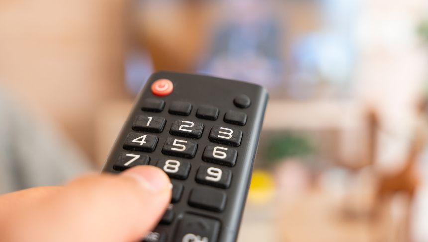 Pilot telewizyjny trzymany w dłoni, w tle nieostry obraz telewizora.