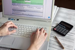 Osoba pracuje przy laptopie, wypełniając formularz podatkowy obok kalkulatora i dokumentów.