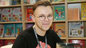 Sławomir Gortych. Młody mężczyzna w okularach, uśmiecha się na tle kolorowych książek. Siedzi przy stole z książkami.