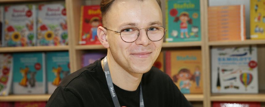 Sławomir Gortych. Młody mężczyzna w okularach, uśmiecha się na tle kolorowych książek. Siedzi przy stole z książkami.