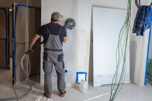 Mężczyzna w roboczym stroju szlifuje ścianę w nowym budynku, otoczenie w trakcie remontu.