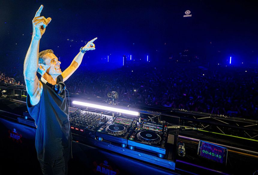 Armin Van Buuren na scenie, uśmiechnięty, unosi ręce w górę, w tle tłum i kolorowe światła.