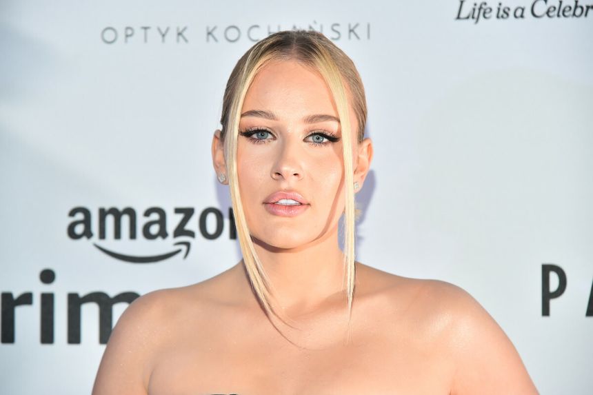 Angelika „Andziaks” Trochonowicz w blond włosach i makijażu, w eleganckiej sukni na tle reklamowym.