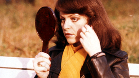 Kobieta w skórzanej kurtce trzyma lusterko i poprawia włosy, na tle jesiennego parku.