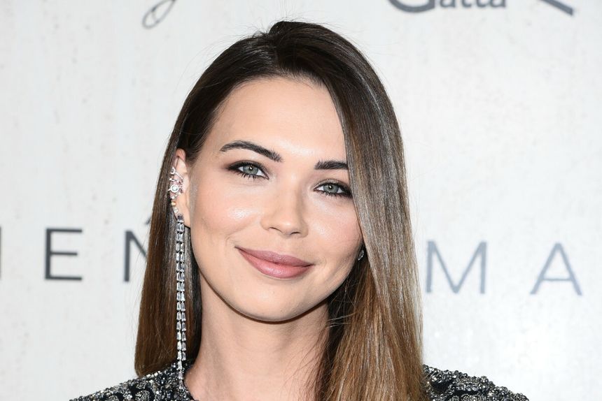 Uśmiechnięta Sandra Kubicka z długimi włosami i elegancką biżuterią, na tle jasnej ściany.