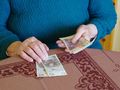 Dwie ręce starszej osoby trzymają banknoty na blacie w kolorze brązowym z wzorem.