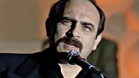 Mężczyzna z wąsem śpiewa do mikrofonu, na tle zamazanego tła.