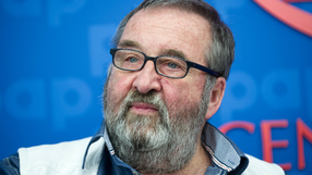 Mężczyzna z siwą brodą, w okularach, uśmiecha się na tle niebieskiego tła.