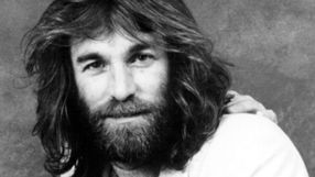 Mężczyzna z długimi włosami i brodą, uśmiechający się do kamery na szarym tle.