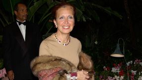 Kobieta w eleganckiej sukience z biżuterią, trzymająca futro w otoczeniu roślin.