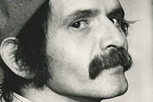 Mężczyzna z wąsem i czapką, spogląda z boku, w tle rozmyte kształty. Czarnobiałe zdjęcie.