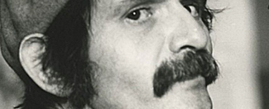 Mężczyzna z wąsem i czapką, spogląda z boku, w tle rozmyte kształty. Czarnobiałe zdjęcie.