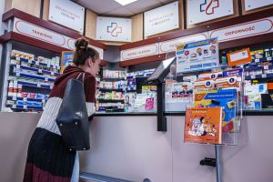 Kobieta stoi przy ladzie w aptece, w tle półki z lekami i materiały informacyjne.