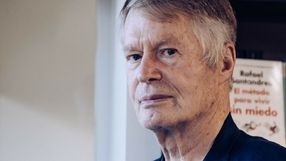 Mężczyzna z siwymi włosami patrzy poważnie w stronę obiektywu. W tle widoczne książki.