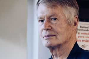 Mężczyzna z siwymi włosami patrzy poważnie w stronę obiektywu. W tle widoczne książki.