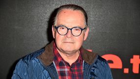 Mężczyzna w okrągłych niebieskich okularach, w niebieskiej kurtce, z uśmiechem na tle czarnej ściany.