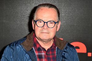 Mężczyzna w okrągłych niebieskich okularach, w niebieskiej kurtce, z uśmiechem na tle czarnej ściany.