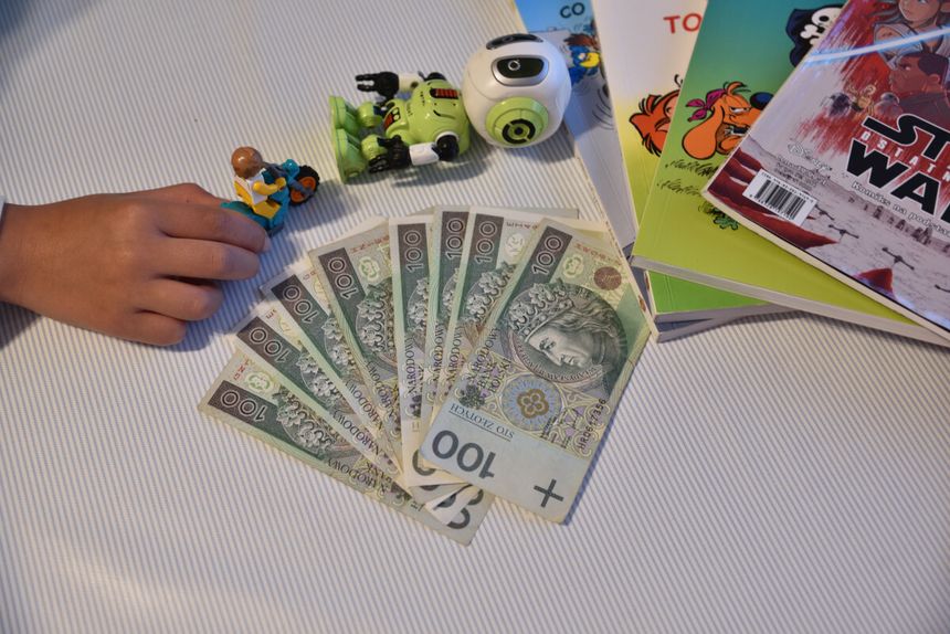 Na zdjęciu widoczna jest ręka dziecka trzymająca figurkę, obok banknoty i książki.