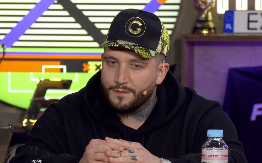 Michał „Boxdek” Baron w czarnej bluzie i czapce z motywem wężowym. Siedzi przy stole z butelką wody.