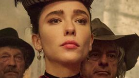 Kobieta w eleganckim stroju, z wyrazem zadumy, w tle mężczyźni w kapeluszach.
