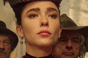 Kobieta w eleganckim stroju, z wyrazem zadumy, w tle mężczyźni w kapeluszach.