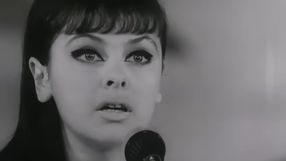 Młoda kobieta z długimi włosami i mocnym makijażem, mówi do mikrofonu. Czarno-białe zdjęcie.