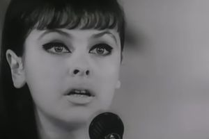 Młoda kobieta z długimi włosami i mocnym makijażem, mówi do mikrofonu. Czarno-białe zdjęcie.