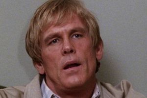 Mężczyzna z krótkimi, jasnymi włosami, siedzi z poważnym wyrazem twarzy.