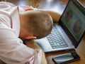Mężczyzna w różowej koszuli opiera głowę na dłoniach, siedząc przy laptopie.