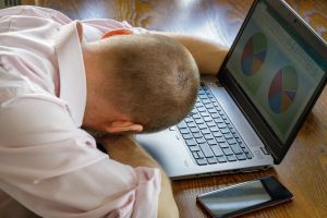 Mężczyzna w różowej koszuli opiera głowę na dłoniach, siedząc przy laptopie.