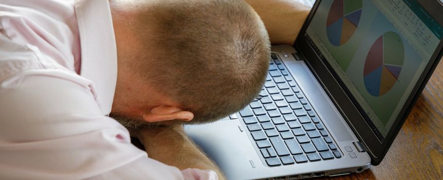 Mężczyzna w różowej koszuli opiera głowę na dłoniach, siedząc przy laptopie.