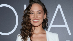 Młoda kobieta z kręconymi włosami uśmiecha się, stojąc na tle ciemnej ściany z napisami.