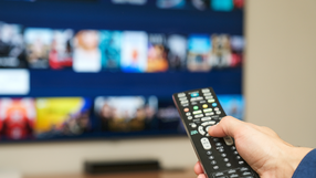 Ręka trzymająca pilot do telewizora, w tle ekran z ikonami filmów i programów.