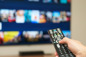 Ręka trzymająca pilot do telewizora, w tle ekran z ikonami filmów i programów.