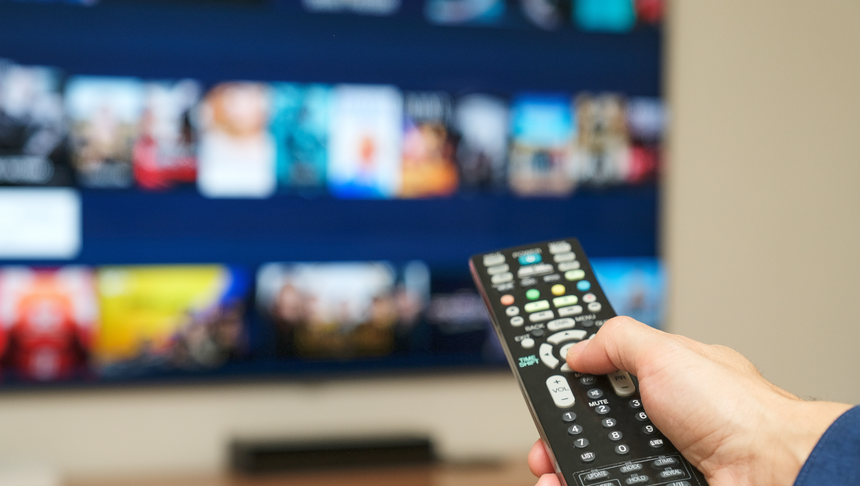 Ręka trzymająca pilot do telewizora, w tle ekran z ikonami filmów i programów.