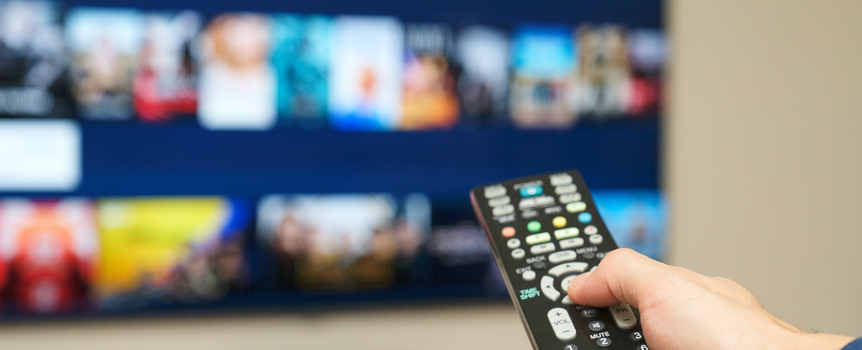 Ręka trzymająca pilot do telewizora, w tle ekran z ikonami filmów i programów.