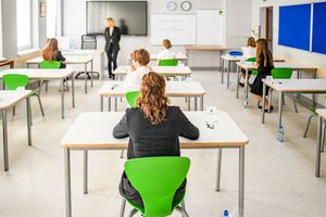 Uczniowie siedzą przy biurkach w klasie na egzaminie, nauczycielka prowadzi zajęcia.
