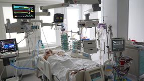 Szpitalna sala z pacjentem na łóżku, otoczonym sprzętem medycznym monitorującym.