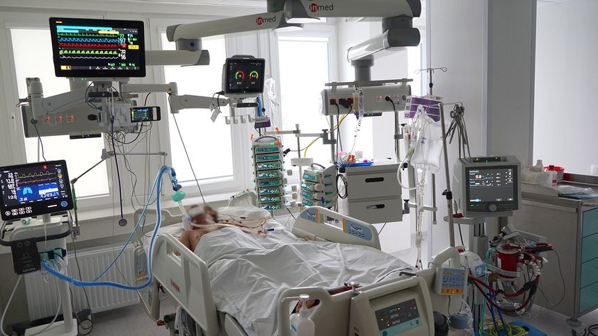 Szpitalna sala z pacjentem na łóżku, otoczonym sprzętem medycznym monitorującym.