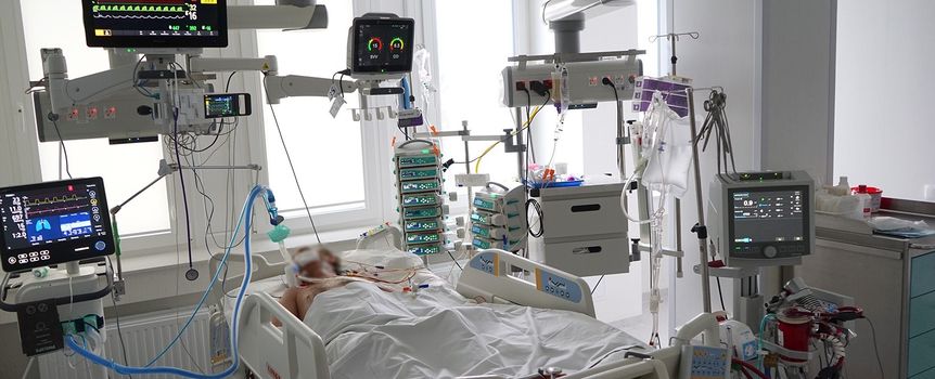Szpitalna sala z pacjentem na łóżku, otoczonym sprzętem medycznym monitorującym.