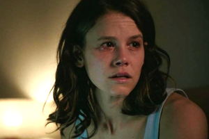 Kobieta w bluzce bez rękawów, z wyraźnym smutkiem i łzami w oczach, siedzi w słabym świetle.